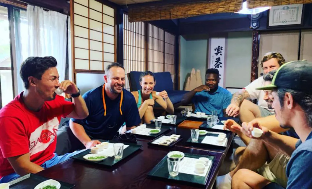 A group of people sit around a wooden table in a traditional Japanese room with tatami mats, enjoying small cups of tea. They are smiling and engaged in conversation. The room features shoji screens and a warm, inviting atmosphere.