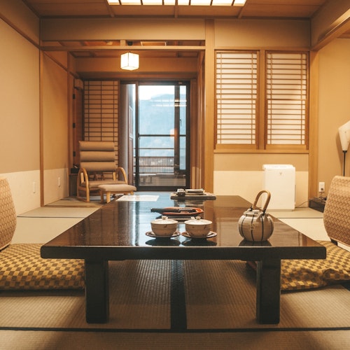 Ryokan A traditional Japanese room with tatami flooring, shoji screens, and a low black wooden table set with tea cups and a teapot. Two cushions are placed on the floor in front of the table. A wooden chair and lamp are visible in the background near large windows.