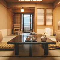 Best Ryokan in Kyoto A traditional Japanese room with tatami flooring, shoji screens, and a low black wooden table set with tea cups and a teapot. Two cushions are placed on the floor in front of the table. A wooden chair and lamp are visible in the background near large windows.