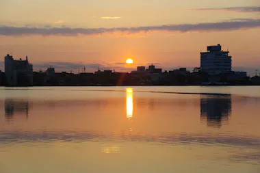 A calm lake reflects the orange and yellow colors of the sunrise, with the sun just above the horizon, silhouetted buildings, and a partly cloudy sky in the background.