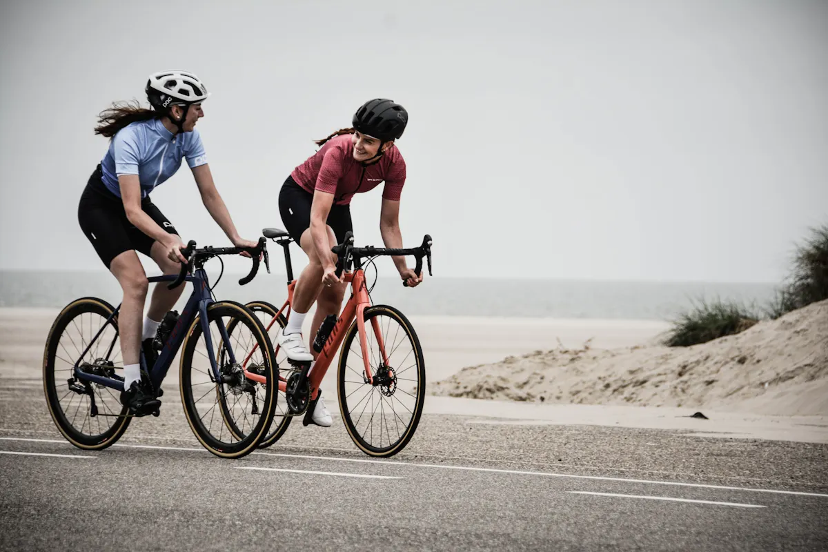 Two cyclists ride side by side on a coastal road, engaging in conversation. The rider on the left wears a blue jersey and black shorts, while the rider on the right wears a pink jersey and black shorts. Both wear helmets, with the sea and sand dunes visible in the background.