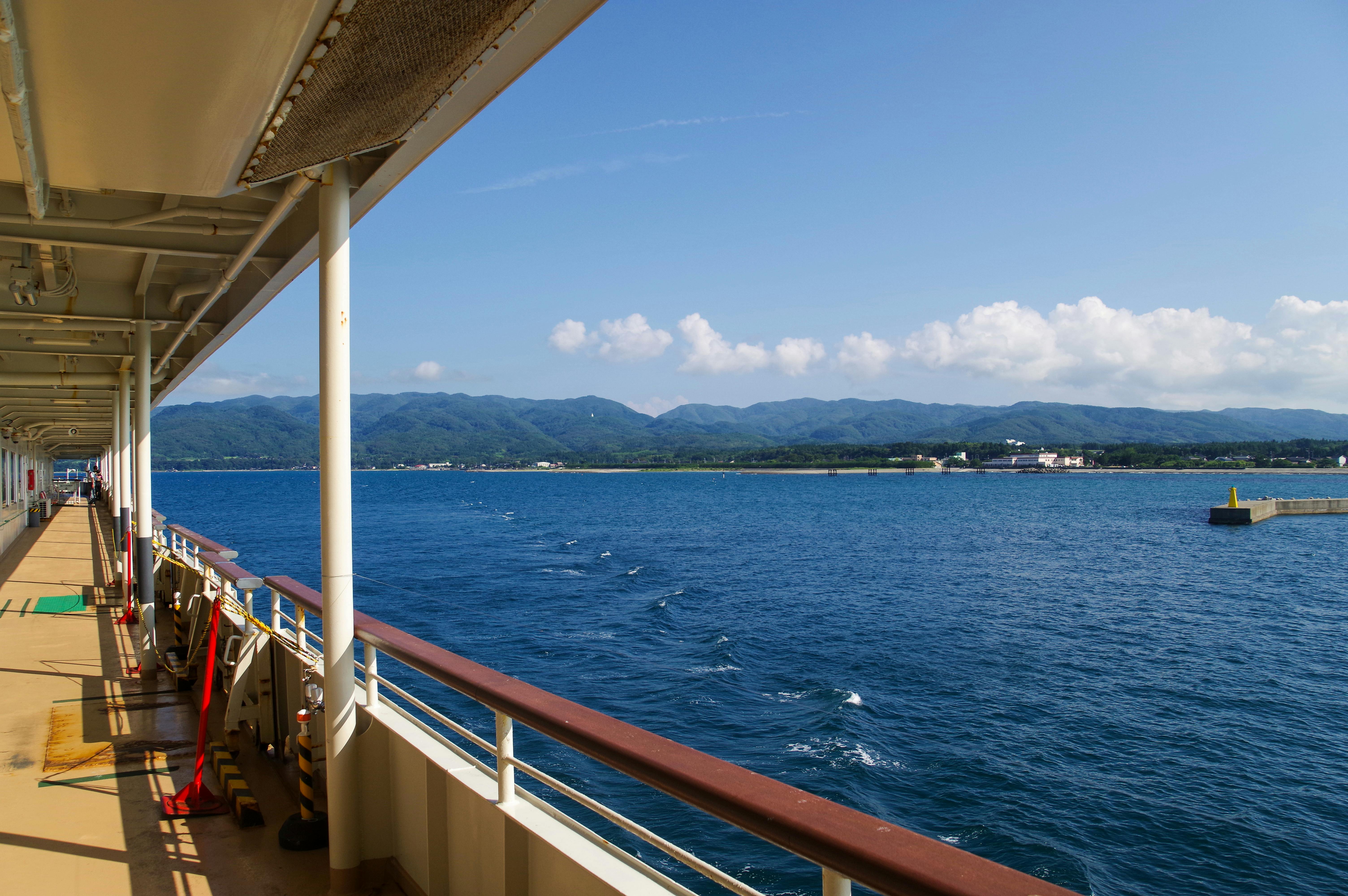 View from a ship’s deck with railings, overlooking calm blue water and a distant shoreline with mountains and buildings under a clear, sunny sky with scattered clouds.