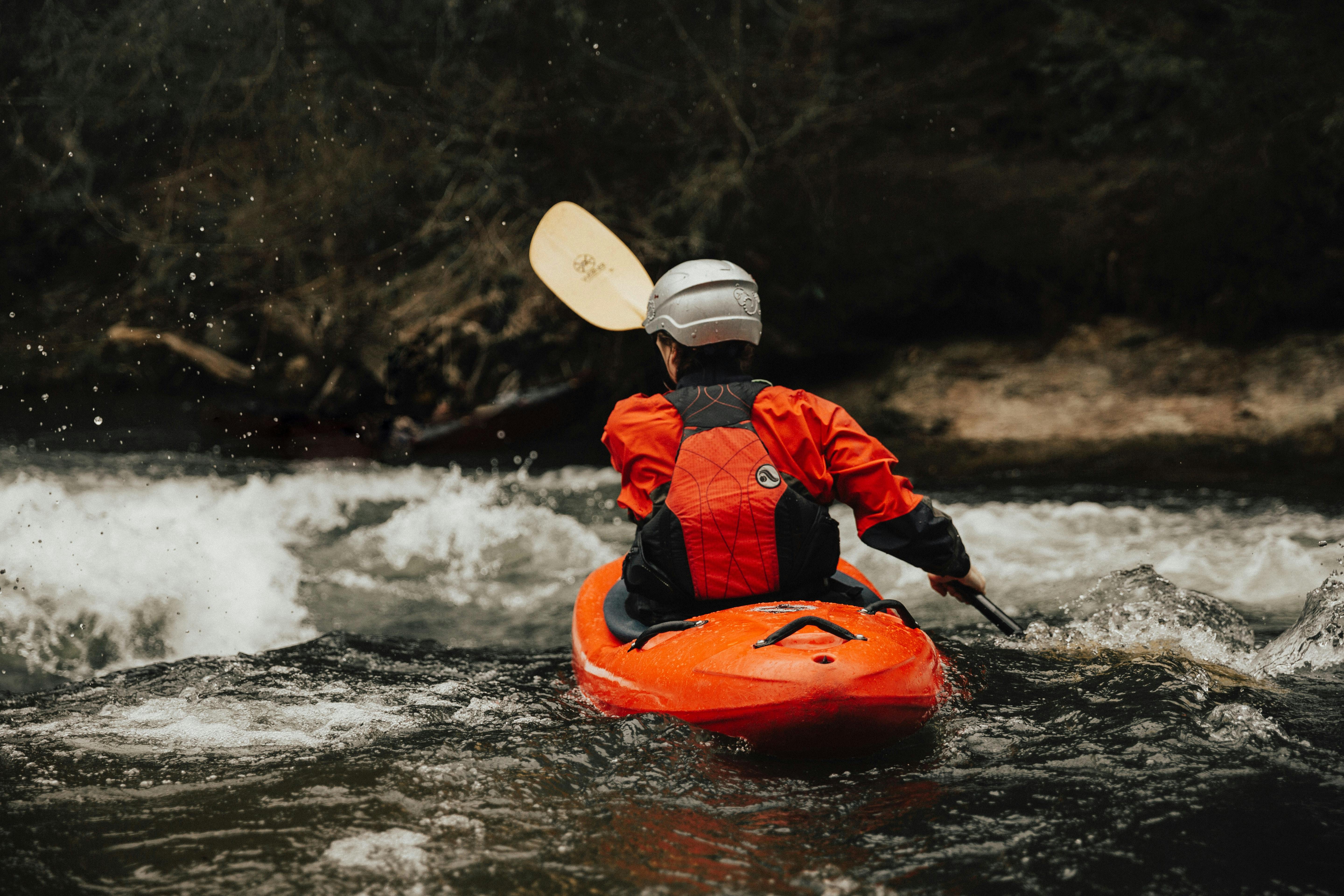 A person in an orange kayak wearing a helmet and life jacket paddles through rough river water, facing away from the camera, surrounded by swirling rapids and dark trees.
