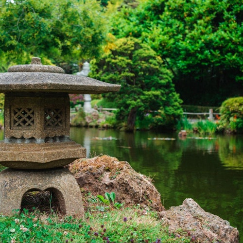 Japanese Garden A serene Japanese garden featuring a stone lantern in the foreground, lush greenery and trees, a pond with calm water, and a bridge in the background. The scene is tranquil and natural.