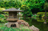 A serene Japanese garden featuring a stone lantern in the foreground, lush greenery and trees, a pond with calm water, and a bridge in the background. The scene is tranquil and natural.