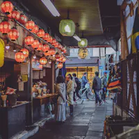 Shimokitazawa A narrow, dimly-lit alley in a city with red and yellow lanterns hanging overhead. People are gathered at a small food stall on the left, with scooters and pedestrians visible in the background. The atmosphere is bustling and lively.