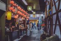 A narrow, dimly-lit alley in a city with red and yellow lanterns hanging overhead. People are gathered at a small food stall on the left, with scooters and pedestrians visible in the background. The atmosphere is bustling and lively.