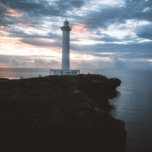 Lighthouse in Okinawa A lone white lighthouse stands on a rocky cliff, overlooking a calm ocean at sunset. The sky is filled with moody clouds, with hints of orange and pink from the setting sun. The dark rocks contrast with the soft light of the evening sky.