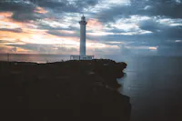 A lone white lighthouse stands on a rocky cliff, overlooking a calm ocean at sunset. The sky is filled with moody clouds, with hints of orange and pink from the setting sun. The dark rocks contrast with the soft light of the evening sky.