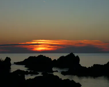A dark, rocky coastline is silhouetted against a calm sea at sunset, with vibrant orange and yellow light illuminating the clouds near the horizon. The sky gradually fades to deep blue as evening approaches.