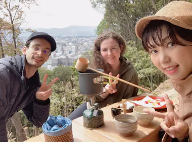 Three people smile and make peace signs while enjoying tea outdoors at a table overlooking a cityscape and trees, with tea utensils and snacks in front of them.