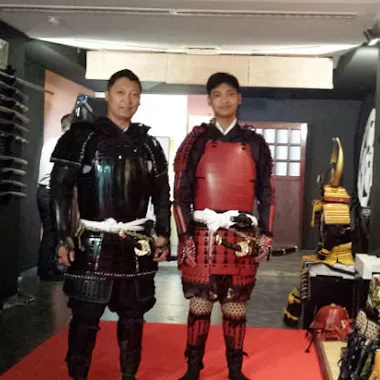 Samurai Armor Two people stand indoors on a red carpet wearing traditional Japanese samurai armor, one in black and one in red. Swords and various helmets are displayed on the walls and racks around them.