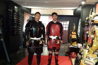 Two people stand indoors on a red carpet wearing traditional Japanese samurai armor, one in black and one in red. Swords and various helmets are displayed on the walls and racks around them.