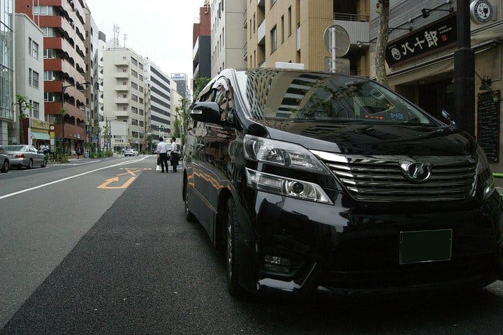 A black minivan is parked on a city street lined with tall buildings. Several people walk on the sidewalk in the background under a cloudy sky. Japanese signs are visible on some storefronts.