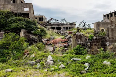 Overgrown plants and grass cover crumbling stone ruins and rubble from abandoned concrete buildings under a cloudy sky, suggesting a once-inhabited, now deserted area.