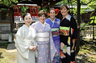 Four women wearing colorful kimonos stand together outdoors in a garden with trees and a traditional Japanese building in the background, smiling at the camera.