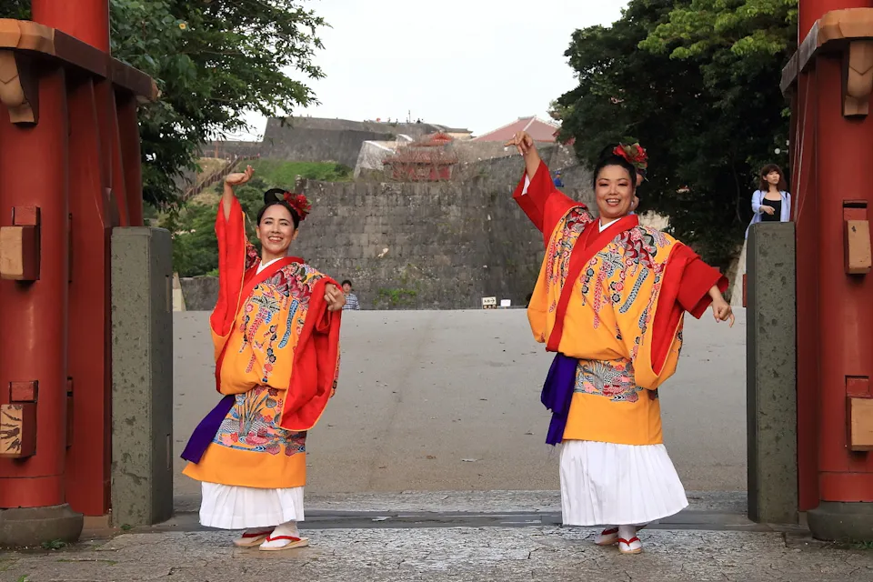 Ryukyuan Traditions 两个微笑的女人身穿鲜艳的传统服饰,在一座历史建筑前表演舞蹈。她们穿着色彩丰富、图案独特的服装,色调包括红色、橙色和白色,站在一个铺砌的区域,周围有巨大的红色柱子,背景中有绿树和石墙。