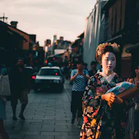 Geisha A person dressed in traditional Japanese attire walks down a busy street lined with buildings. The person is wearing a floral-patterned kimono and elaborate headpiece. Other people and a car are seen in the background during sunset.