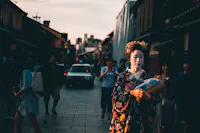 A person dressed in traditional Japanese attire walks down a busy street lined with buildings. The person is wearing a floral-patterned kimono and elaborate headpiece. Other people and a car are seen in the background during sunset.