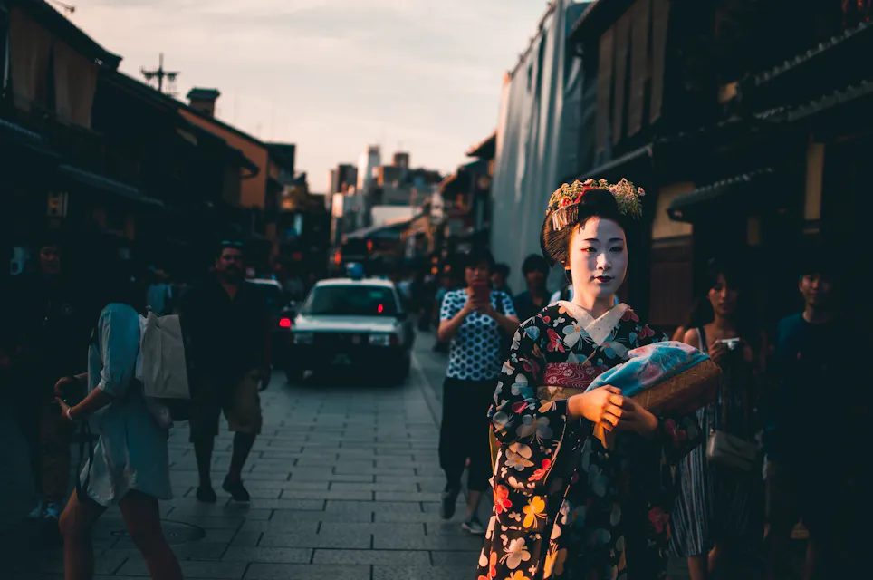 A person dressed in traditional Japanese attire walks down a busy street lined with buildings. The person is wearing a floral-patterned kimono and elaborate headpiece. Other people and a car are seen in the background during sunset.