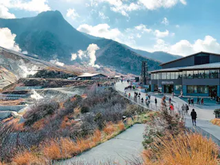 People walk along a paved path near modern buildings and steaming hot springs in a mountainous area on a sunny day, with hills and clouds in the background.