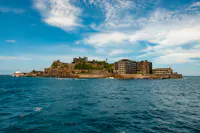 An abandoned island with ruined concrete buildings surrounded by blue ocean under a partly cloudy sky. The island's rocky land and derelict structures are prominent against the water.