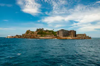 An abandoned island with ruined concrete buildings surrounded by blue ocean under a partly cloudy sky. The island's rocky land and derelict structures are prominent against the water.