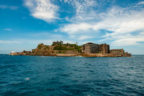 An abandoned island with ruined concrete buildings surrounded by blue ocean under a partly cloudy sky. The island's rocky land and derelict structures are prominent against the water.