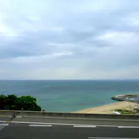 Awaji Island View from a road of a calm, vast ocean with a slightly cloudy sky above. On the right, a curved breakwater and marina with several small boats are visible. Green foliage and sandy beach edge the road in the foreground.