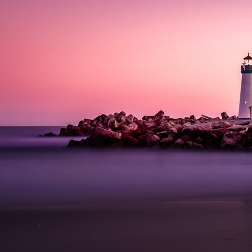 Shimonoseki Lighthouse A lighthouse stands at the end of a rocky pier, illuminated against a serene sea at dusk. The sky is a gradient of purples and pinks, casting a tranquil ambiance over the calm waters and shoreline.