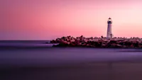 A lighthouse stands at the end of a rocky pier, illuminated against a serene sea at dusk. The sky is a gradient of purples and pinks, casting a tranquil ambiance over the calm waters and shoreline.