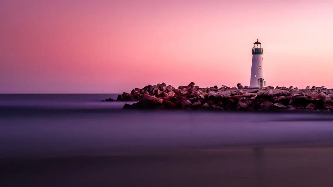 A lighthouse stands at the end of a rocky pier, illuminated against a serene sea at dusk. The sky is a gradient of purples and pinks, casting a tranquil ambiance over the calm waters and shoreline.