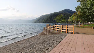 A scenic lakeside path with a wooden fence, stone steps leading to the water, and tree-covered hills in the background under a partly cloudy sky.