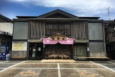 A small theater building with a traditional design, featuring a mural of an audience under a pink awning. Glass panels and wooden slats decorate the facade. Two parking spaces with painted wheelchair symbols are in the foreground.