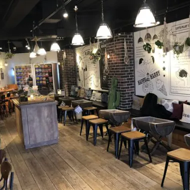 Hedgehog Cafe A cozy cafe interior with wooden floors, small tables and chairs, cushioned bench seating, hanging plants, and wall art featuring illustrations and text. Warm pendant lights create a welcoming atmosphere.