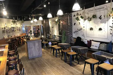 A cozy cafe interior with wooden floors, small tables and chairs, cushioned bench seating, hanging plants, and wall art featuring illustrations and text. Warm pendant lights create a welcoming atmosphere.