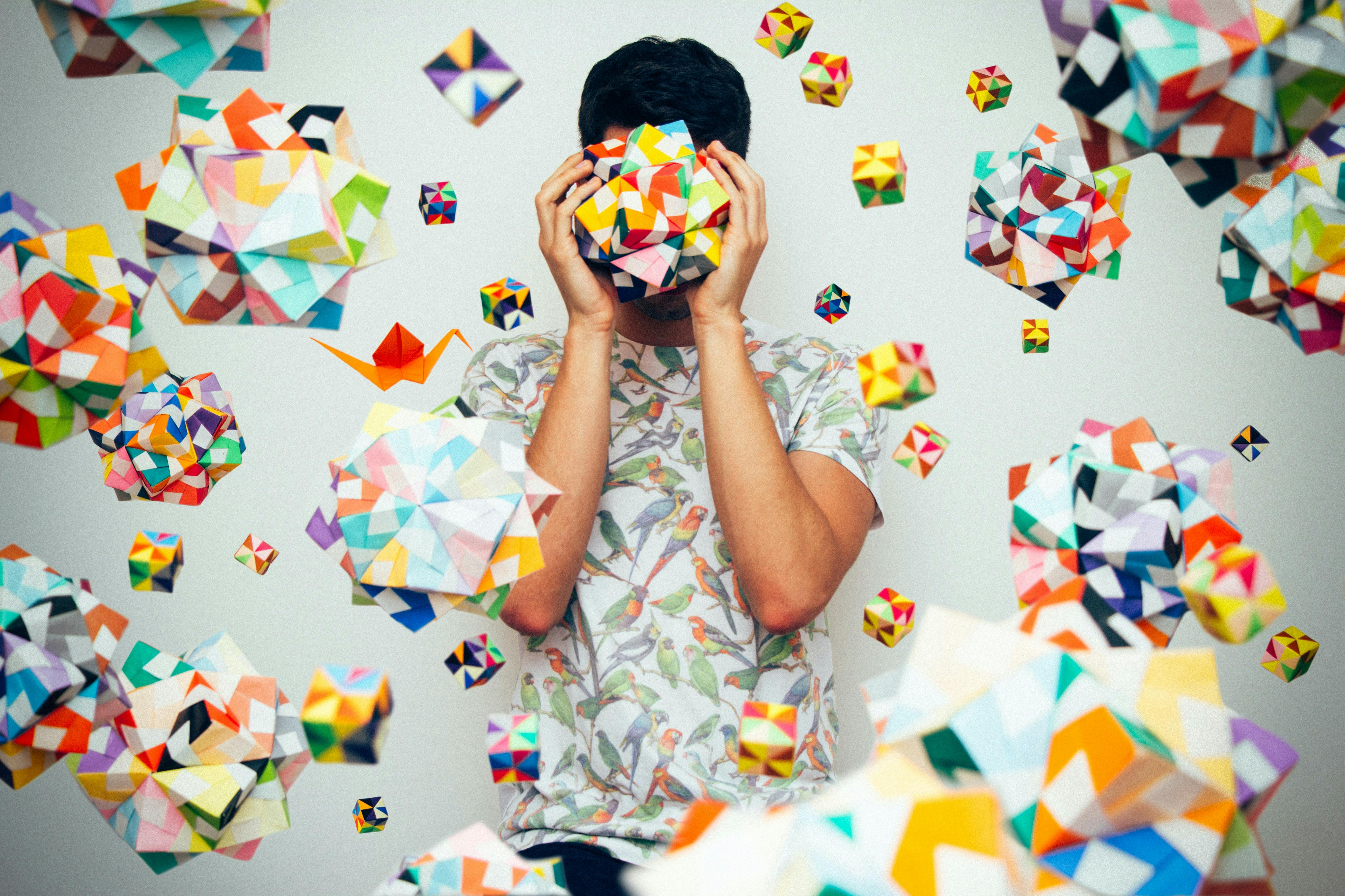 A person in a patterned shirt covers their face with a colorful geometric shape, surrounded by floating, multifaceted, multicolored cubes against a plain background.