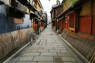 A narrow, stone-paved alley lined with traditional wooden buildings in Japan; a bicycle is parked on the left, and bamboo fencing frames both sides under a bright sky.