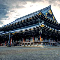 Higashi Honganji Temple A traditional Japanese temple with intricate wooden architecture and a dark roof sits against a dramatic cloudy sky. The temple features ornate gold and black details, a raised platform, and multiple levels with curving eaves. The foreground shows a gravel courtyard.