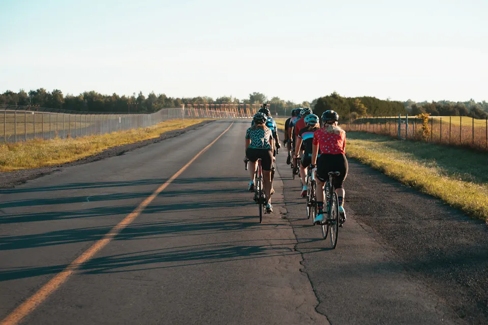 A group of cyclists riding on a paved road. The road is surrounded by grassy fields and lined with a wire fence on the right side. The sky is clear, suggesting a sunny day. The cyclists are wearing helmets and colorful cycling attire, pedaling in a close formation.
