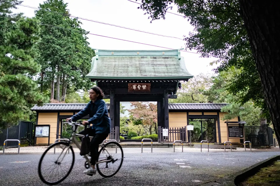 A person rides a bicycle past a traditional Japanese gate surrounded by lush greenery. The gate has a green roof and a wooden sign with Japanese characters. The scene is framed by trees on either side, and the ground is speckled with sunlight filtering through the branches.
