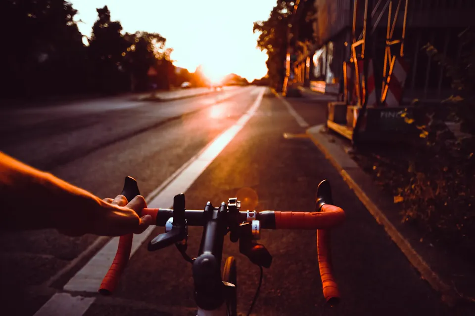 Close-up of a person riding a bicycle on a road at sunset. The handlebars are wrapped in red tape, and the rider's left hand grips one side. The sun is low on the horizon, casting a warm glow on the scene. Trees and buildings line the road.