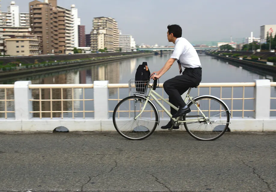 A man in business attire rides a bicycle across a bridge over a river on an overcast day. High-rise buildings line both sides of the river in the background. The man has a black bag in the bicycle's front basket.