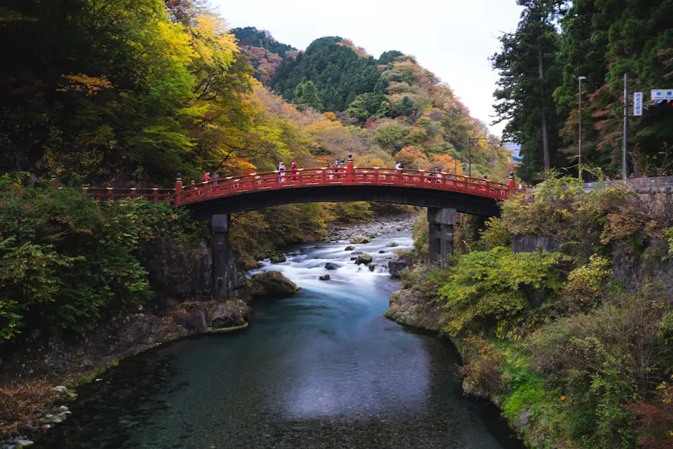 Nikko 一座风景如画的红色桥梁横跨宁静的河流,周围环绕着郁郁葱葱的绿叶和秋色树木。人们可以看到在桥上行走,享受美丽的景色。河水缓缓流过,河岸边有岩石和绿意盎然的植物。
