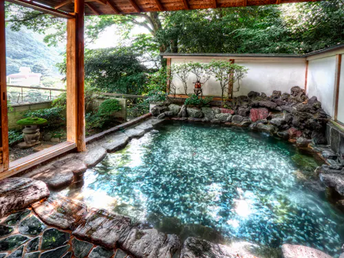 Hakone Onsen A serene outdoor hot spring bath surrounded by natural rocks and lush greenery. The water is clear and reflective, and a small traditional lantern adds ambiance. A wooden opening offers a view of the surrounding landscape.