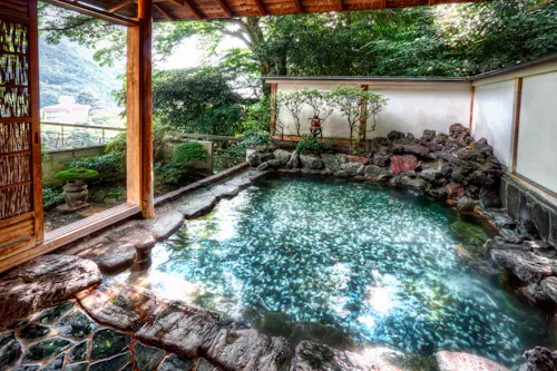 A serene outdoor hot spring bath surrounded by natural rocks and lush greenery. The water is clear and reflective, and a small traditional lantern adds ambiance. A wooden opening offers a view of the surrounding landscape.