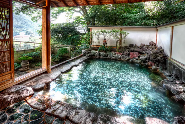 A serene outdoor hot spring bath surrounded by natural rocks and lush greenery. The water is clear and reflective, and a small traditional lantern adds ambiance. A wooden opening offers a view of the surrounding landscape.