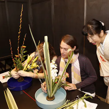 Ikebana Workshop Two people are arranging flowers in blue vases at a table while a woman in a white and yellow kimono observes and smiles. The scene appears to be an ikebana (Japanese flower arranging) class.