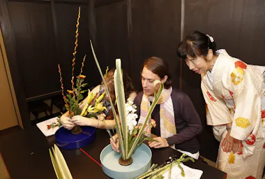 Two people are arranging flowers in blue vases at a table while a woman in a white and yellow kimono observes and smiles. The scene appears to be an ikebana (Japanese flower arranging) class.