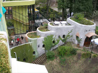 Aerial view of the Studio Ghibli Museum in Mitaka, Japan. The architecture features organic, curved buildings with a green roof and ivy-covered walls. Visitors are walking around the museum's exterior, and a garden area is visible in the foreground.